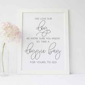 May include: A framed print with the text "WE LOVE OUR dog, AS WE'RE SURE YOU KNOW. SO TAKE A doggie bag FOR YOURS...TO GO!" in black script. A vase with pink flowers is to the left.