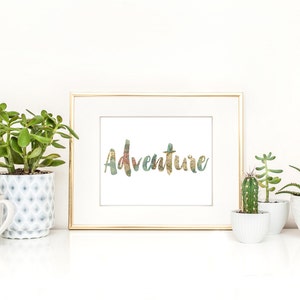 May include: A framed print with the word "Adventure" in a colorful, textured font. The gold-framed artwork is displayed on a white surface, flanked by various potted succulents and cacti in white and patterned ceramic pots, creating a bright, inviting scene.