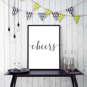 May include: A framed white print with the word "cheers" in elegant black script. The print is displayed on a black table with decorative bottles, glasses, and a candle holder. A string of decorative pennants hangs above.