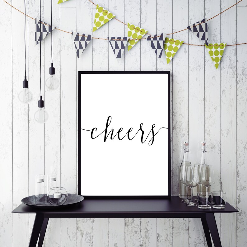 May include: A framed white print with the word "cheers" in elegant black script. The print is displayed on a black table with decorative bottles, glasses, and a candle holder. A string of decorative pennants hangs above.