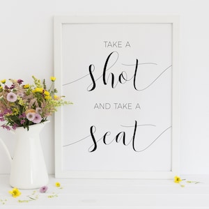 May include: A framed print with the words "TAKE A shot AND TAKE A seat" in elegant black script. A white pitcher filled with colorful flowers sits to the left. Yellow and purple flowers are scattered on the white surface.