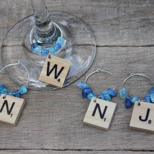May include: Wine glass charms with Scrabble tile letters and blue gemstone beads. Each charm has a silver hoop and a wooden tile with a letter and number. The charms are on a wooden surface.