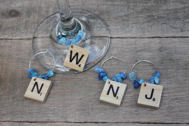 May include: Wine glass charms with Scrabble tile letters and blue gemstone beads. Each charm has a silver hoop and a wooden tile with a letter and number. The charms are on a wooden surface.