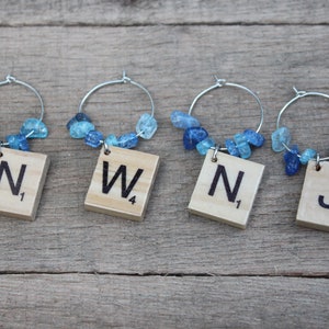 May include: Four wine charms with silver hoops, each featuring a wooden Scrabble tile with a letter and number. The charms are adorned with blue gemstone beads. The letters are N, W, N, and J.