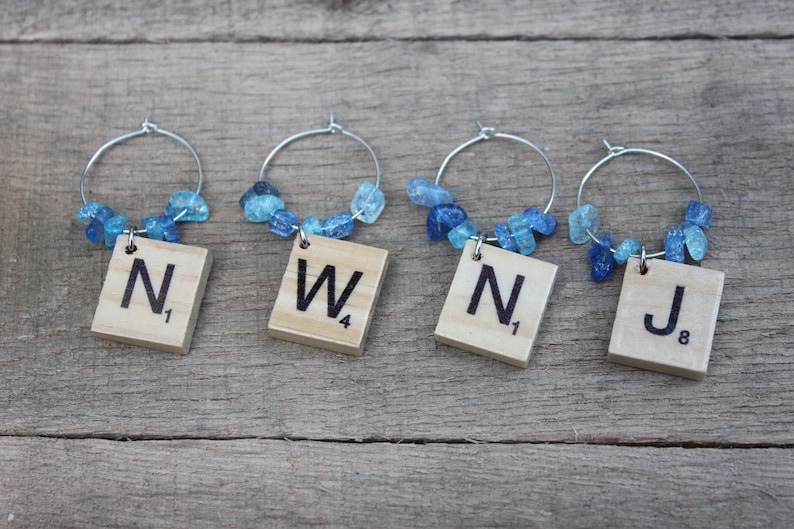 May include: Four wine charms with silver hoops, each featuring a wooden Scrabble tile with a letter and number. The charms are adorned with blue gemstone beads. The letters are N, W, N, and J.