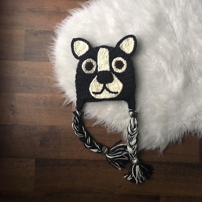 May include: A handmade black and white crochet hat shaped like a dog's face. The hat features a white muzzle, ears, and accents, with brown eyes. Braided black and white tassels hang from the ear flaps. The hat is displayed on a white fluffy surface.