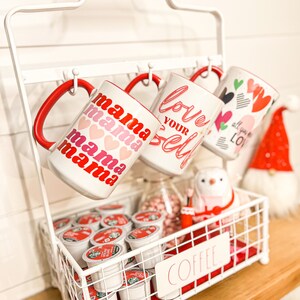 Pu&ograve; includere: Un porta tazze in metallo bianco con manico rosso che tiene quattro tazze con disegni a tema San Valentino. Le tazze dicono "mama mama mama", "love your self" e "all you need is love". Un cestino di filo bianco con la parola "coffee" sul davanti contiene una variet&agrave; di capsule di caff&egrave;.