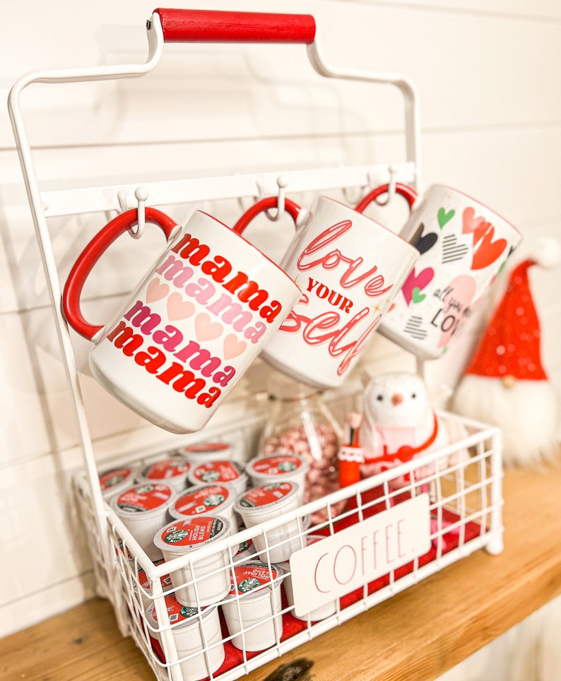 Pu&ograve; includere: Un porta tazze in metallo bianco con manico rosso che tiene quattro tazze con disegni a tema San Valentino. Le tazze dicono "mama mama mama", "love your self" e "all you need is love". Un cestino di filo bianco con la parola "coffee" sul davanti contiene una variet&agrave; di capsule di caff&egrave;.