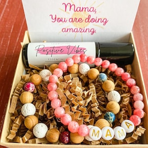 May include: A gift box filled with brown paper shreds, a pink essential oil roller bottle labeled "Positive Vibes Essential", a pink beaded bracelet with a "MAMA" charm, and a card that reads "Mama, you are doing amazing."