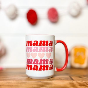 Pu&ograve; includere: Tazza in ceramica bianca con bordo e manico rossi. La tazza ha le parole "mama mama mama mama" stampate su di essa in un font rosso e rosa sfumato con cuori rosa tra ogni parola.