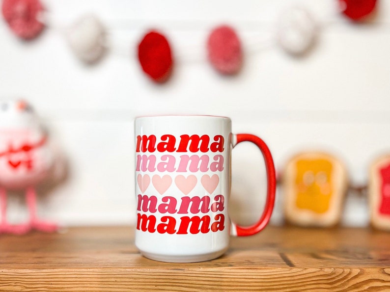 Pu&ograve; includere: Tazza in ceramica bianca con bordo e manico rossi. La tazza ha le parole "mama mama mama mama" stampate su di essa in un font rosso e rosa sfumato con cuori rosa tra ogni parola.