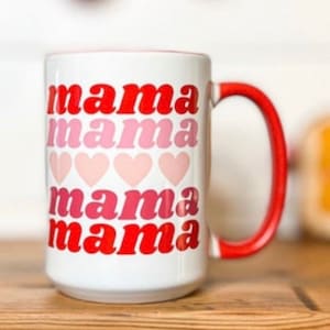 Pu&ograve; includere: Una tazza in ceramica bianca con un manico rosso. La tazza ha le parole "mama mama mama mama" stampate su di essa in lettere rosse e rosa con cuori rosa tra ogni parola.