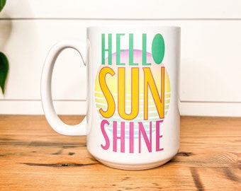 May include: White ceramic mug with the words "HELLO SUN SHINE" in green, yellow, and pink. The design features a colorful sun graphic. The mug has a large handle and is on a wooden surface.