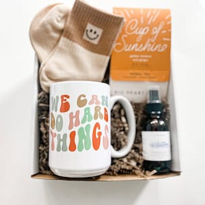 May include: A gift box filled with self-care items, including a white mug with the text "We Can Do Hard Things" in colorful retro font, a pair of beige socks with a smiley face, a small bottle of essential oil, and a box of herbal tea labeled "Cup of Sunshine".