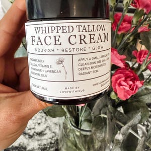 May include: A jar of whipped tallow face cream with a black lid. The label reads "WHIPPED TALLOW FACE CREAM" with details about ingredients and usage. The jar is held in front of a blurred background of pink flowers.