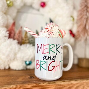 May include: White ceramic mug with a red, green, and black "Merry and Bright" design. The mug is filled with whipped cream and topped with candy canes.