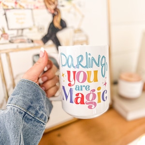 May include: White ceramic mug with the text "Darling you are Magic" in rainbow colors. The text is in a whimsical font.