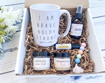 May include: A gift box filled with self-care items. Includes a white mug with "I AM BRAVE BOLD & STRONG" text, a spray bottle, a rollerball, Epsom salt, magnesium balm, and a beaded keychain. The box is filled with brown crinkle paper.