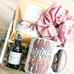 May include: A white ceramic mug with the text "Good Things Take Time" in a retro font with brown, orange, and pink colors. The mug is in a gift box with a pink hair scrunchie, a brown paper bag, and a clear glass bottle with a spray nozzle.