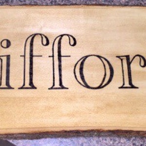 May include: A wooden sign with the name "Clifford" in black, stylized lettering. The sign has a natural wood grain background and a decorative pinecone and branch design in the upper right corner. The edges of the sign are rough and unfinished.