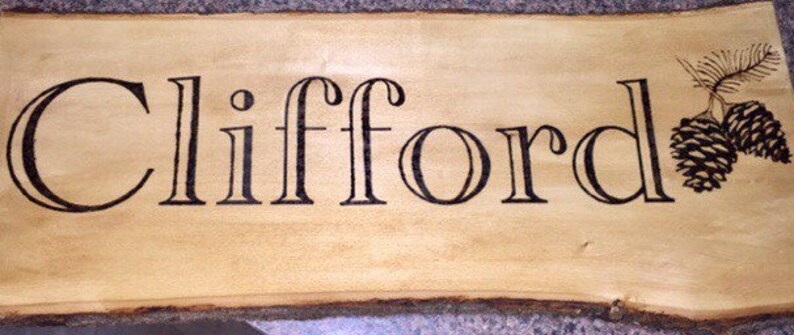May include: A wooden sign with the name "Clifford" in black, stylized lettering. The sign has a natural wood grain background and a decorative pinecone and branch design in the upper right corner. The edges of the sign are rough and unfinished.