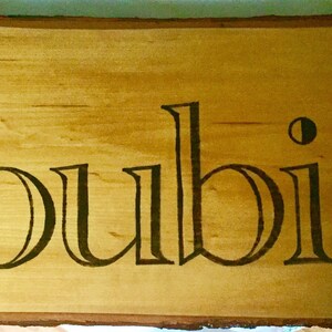 May include: A wooden sign with the name "Loubier" in a decorative font. The text is dark brown and stands out against the light brown wood grain. The sign has a natural, unfinished edge.