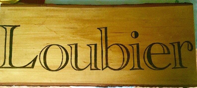 May include: A wooden sign with the name "Loubier" in a decorative font. The text is dark brown and stands out against the light brown wood grain. The sign has a natural, unfinished edge.