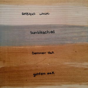 May include: A wooden surface displays various wood stain colors. The samples include "Antique Oak," "Antique White," "Sunbleached," "Summer Oak," "Golden Oak," and "Unstained," each labeled with handwritten text. The wood grain is visible.