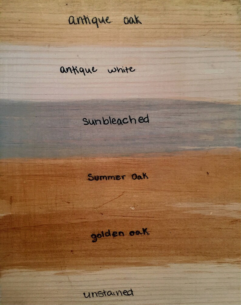 May include: A wooden surface displays various wood stain colors. The samples include "Antique Oak," "Antique White," "Sunbleached," "Summer Oak," "Golden Oak," and "Unstained," each labeled with handwritten text. The wood grain is visible.