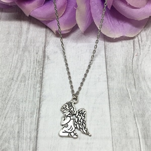May include: Silver-toned necklace featuring a kneeling angel pendant with detailed wings. The angel is praying, and the chain is delicate. The background includes a purple flower and a light gray wooden surface.