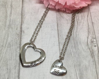 May include: Two silver heart necklaces with the words "Big Sis" and "Little Sis" engraved on them. The necklaces are on a light wood background with a pink flower.