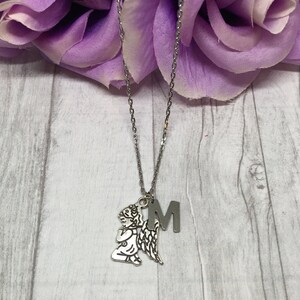 May include: Silver-toned necklace featuring a pendant of a kneeling angel with outstretched wings and the letter "M". The chain is delicate, and the pendants are detailed. The necklace is displayed against a backdrop of purple flowers and a light wood-grain surface.