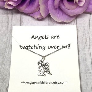 May include: A silver-toned angel pendant necklace on a white card with the text "Angels are watching over me." The card is set against a backdrop of purple flowers and a light wood-grain surface. The Etsy shop URL is also visible.