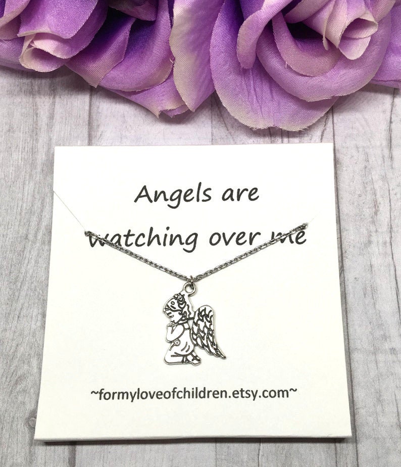 May include: A silver-toned angel pendant necklace on a white card with the text "Angels are watching over me." The card is set against a backdrop of purple flowers and a light wood-grain surface. The Etsy shop URL is also visible.