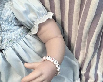 May include: A close-up of a baby doll wearing a light blue dress with white embroidery. The doll is wearing a beaded bracelet that says "I love you" with a red heart bead.