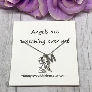May include: A silver-toned necklace with a praying angel pendant and an initial "M" charm. The necklace is displayed on a white card with the text "Angels are watching over me". Purple flowers are in the background.