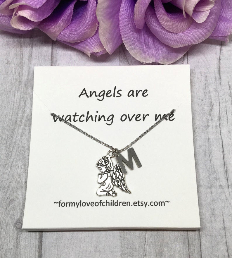 May include: A silver-toned necklace with a praying angel pendant and an initial "M" charm. The necklace is displayed on a white card with the text "Angels are watching over me". Purple flowers are in the background.