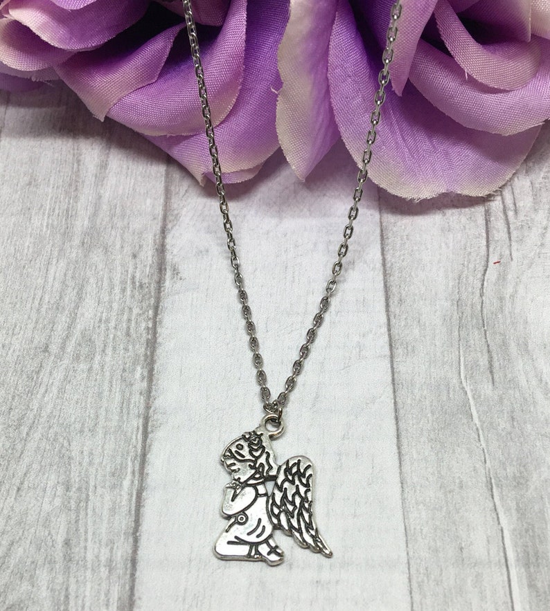 May include: Silver-toned necklace featuring a kneeling angel pendant with detailed wings. The angel is praying, and the chain is delicate. The background includes a purple flower and a light gray wooden surface.