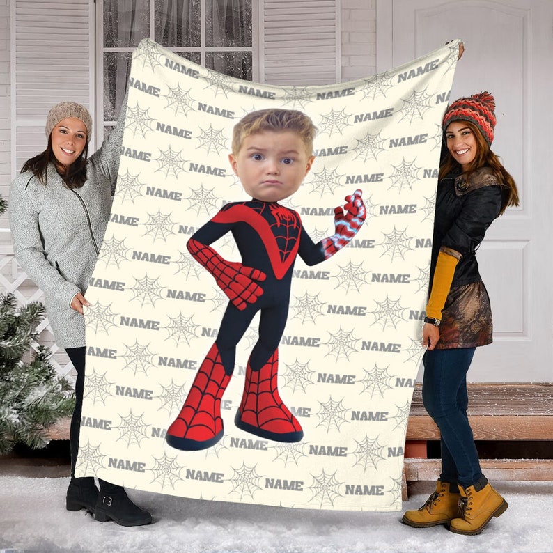 May include: A rectangular throw blanket featuring a repeating pattern of grey spiderwebs and the word NAME in grey text. Centered on the blanket is a large graphic of a child's face on a superhero body wearing a black and red suit.