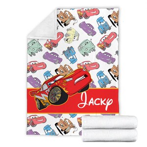 Pu&ograve; includere: Una coperta in pile bianca con un motivo ripetuto di auto dei cartoni animati in rosso, blu, viola, verde, giallo e marrone. Una grande auto da corsa rossa &egrave; centrata sopra una fascia orizzontale rossa con il nome Jacky scritto in bianco.