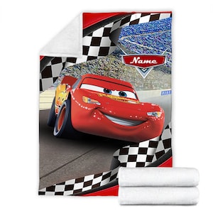 May include: A plush fleece blanket featuring a red race car character on a racetrack with a black and white checkered border. The design includes a stadium background and a red emblem with the word Name in white text. The blanket is folded.