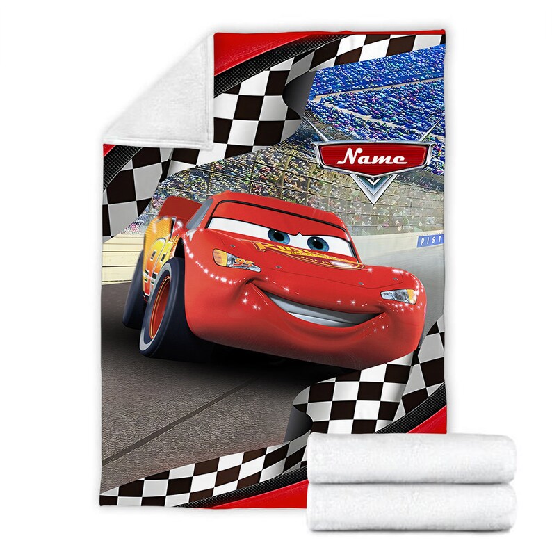 May include: A plush fleece blanket featuring a red race car character on a racetrack with a black and white checkered border. The design includes a stadium background and a red emblem with the word Name in white text. The blanket is folded.