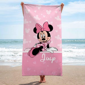 Puede incluir: Una toalla de playa rectangular con fondo rosa y un patr&oacute;n repetitivo de siluetas de Minnie Mouse y estrellas blancas. El centro muestra un gr&aacute;fico de Minnie Mouse con un lazo rosa y el nombre Juip escrito en letra cursiva de color blanco.