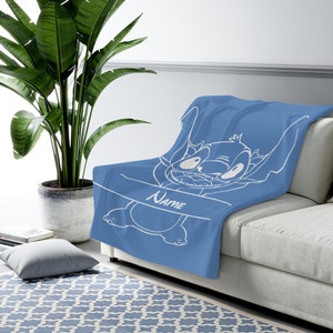 May include: A blue throw blanket featuring a white line art illustration of the character Stitch from Lilo and Stitch. The design includes a horizontal banner across the center with the word Name printed in a white script font on the blanket.