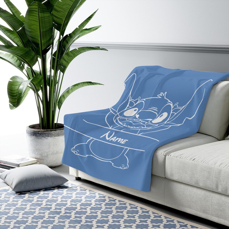 May include: A blue throw blanket featuring a white line art illustration of the character Stitch from Lilo and Stitch. The design includes a horizontal banner across the center with the word Name printed in a white script font on the blanket.