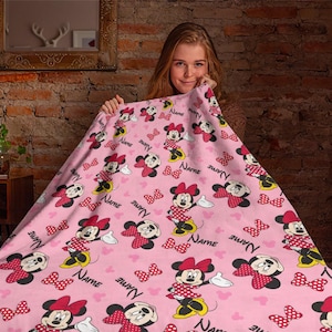 Puede incluir: Manta rosa con un patr&oacute;n repetido de Minnie Mouse, lazos y la palabra "NAME" en negro. Minnie Mouse se representa con su cl&aacute;sico vestido de lunares rojos y blancos y su lazo. La manta es suave y acogedora.