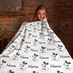 May include: A white fleece blanket featuring a repeating pattern of black Mickey Mouse illustrations and the word Name printed in a black script font. The blanket has a soft texture and displays the character in various standing poses.