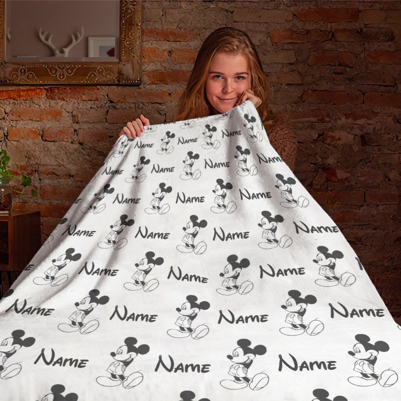 May include: A white fleece blanket featuring a repeating pattern of black Mickey Mouse illustrations and the word Name printed in a black script font. The blanket has a soft texture and displays the character in various standing poses.