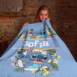 May include: A light blue fleece blanket featuring the character Stitch from Lilo and Stitch, surrounded by tropical flowers and green leaves. The name Sofia is printed in white letters across the center, with additional illustrations of Stitch.
