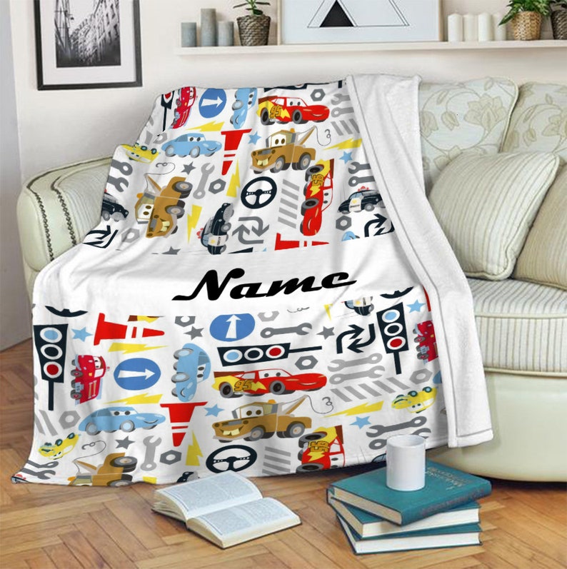 May include: A white fleece blanket featuring a repeating pattern of cartoon cars, tow trucks, traffic lights, road signs, wrenches, and lightning bolts. The centre of the blanket displays the word Name in a bold, black, cursive script font.
